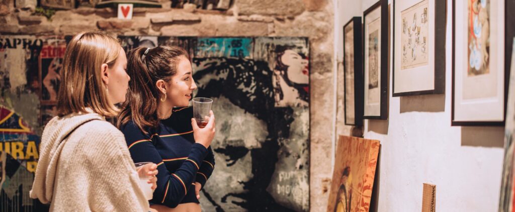 Two young ladies thoughtfully look at a piece of art in a gallery with exposed stone walls, one holds a glass of wine pensively